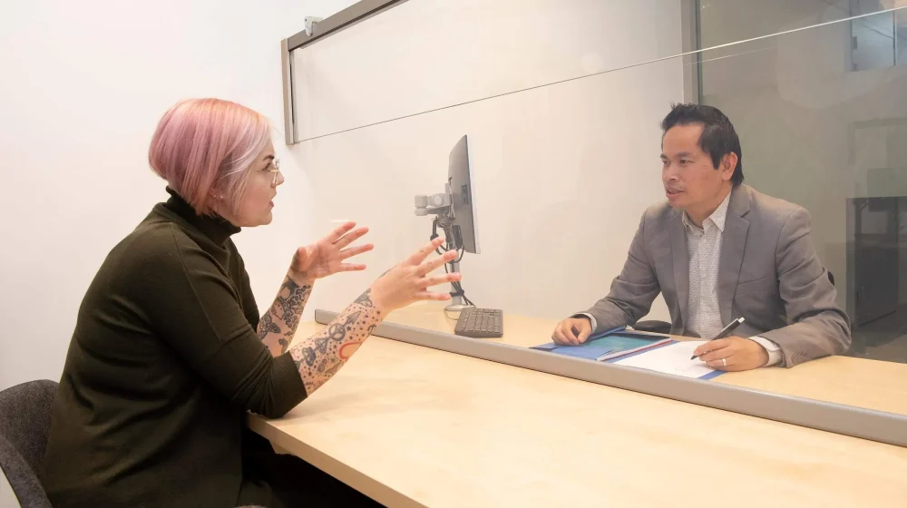Vrouw met roze haar en tatoeages in gesprek met mannelijke medewerker achter balie